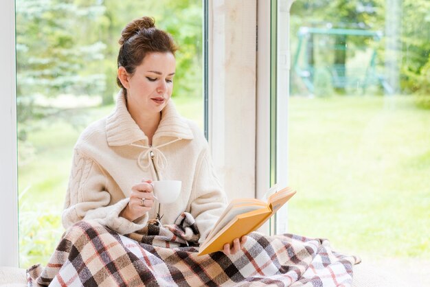 Brunette vrouw in warme trui leesboek met mok koffie zittend op kamer