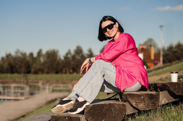 Brunette vrouw in zonnebril en roze shirt zitten en ontspannen in het park over de blauwe hemelachtergrond