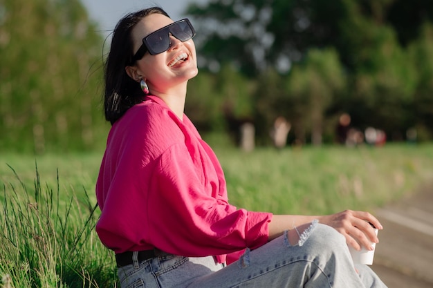 Brunette vrouw in zonnebril zittend op de oever van het meer en chillen in de zonnestralen buiten