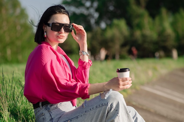 Brunette vrouw in zonnebril zittend op de oever van het meer en chillen in de zonnestralen buiten