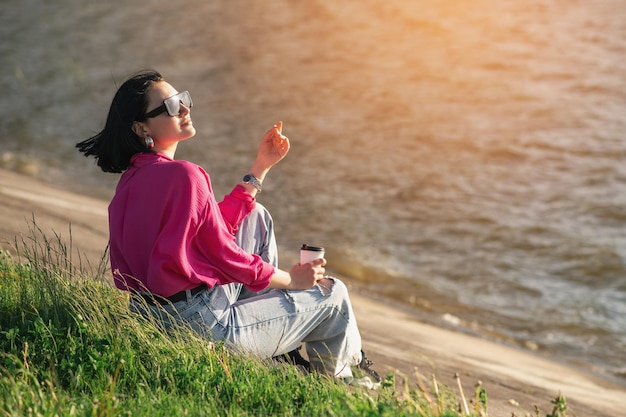 Brunette vrouw in zonnebril zittend op de oever van het meer en chillen in de zonnestralen buiten