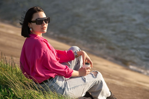 Brunette vrouw in zonnebril zittend op de oever van het meer en chillen in de zonnestralen buiten