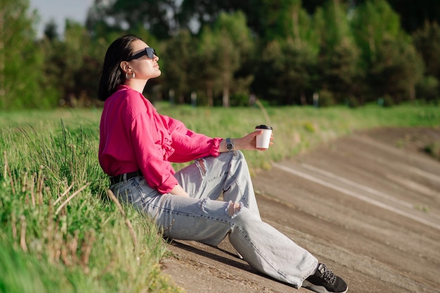 Brunette vrouw in zonnebril zittend op de oever van het meer en chillen in de zonnestralen buiten