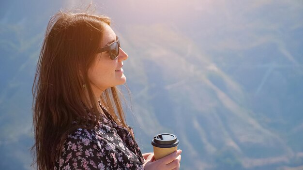 Brunette vrouw met stijlvolle zonnebril in kleur jurk drinkt koffie genietend van prachtig landschap van heuvelachtige vallei in hoogland op zonnige dag close-up