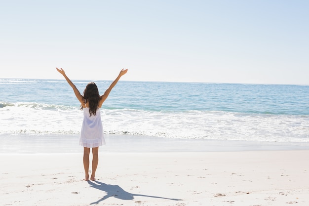 Brunette in white sun dress standing by the water with arms raised