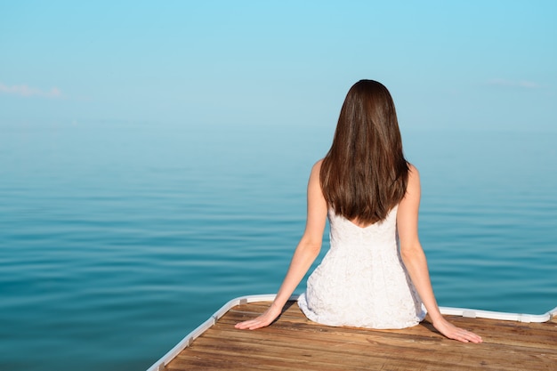 Brunette in white sundress sitting on the pier and looking at the horizontal