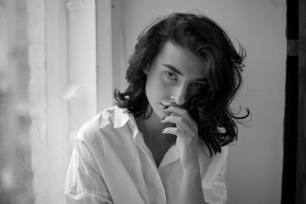 Brunette with curly hair wearing a white shirt posing sitting on the window black and white photo