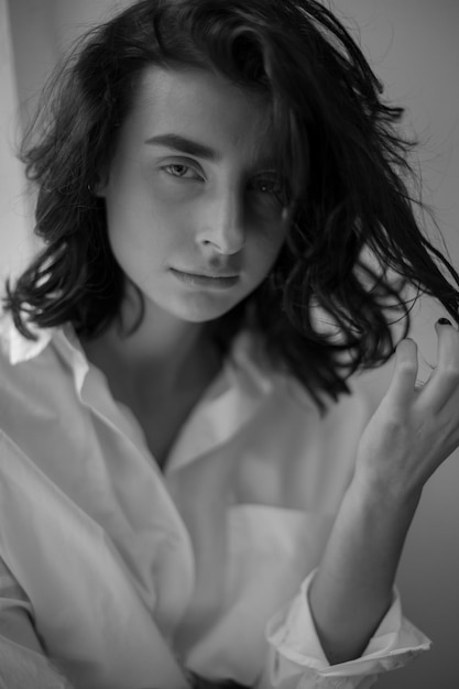 Brunette with curly hair wearing a white shirt posing sitting on the window black and white photo