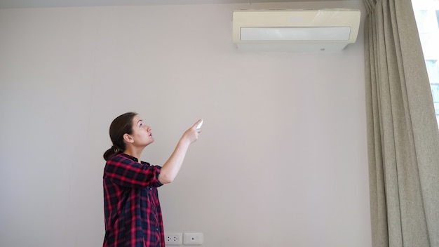 Brunette with long hair in ponytail turns on air conditioner\
with remote control and smiles standing near white wall