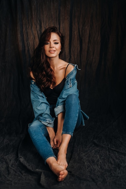 A brunette with long hair poses in a studio sitting on the floor on a black background.