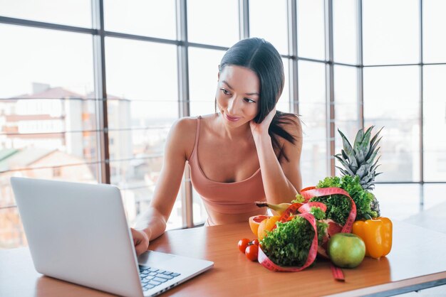 Brunette with slim body type sits by the table with laptop and healthy food.