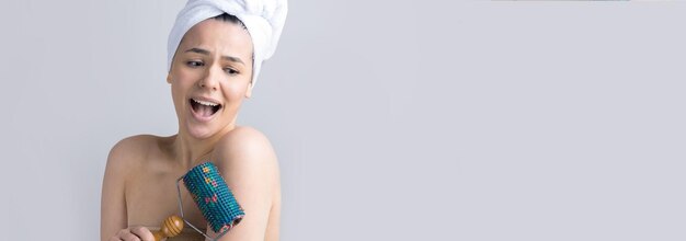 Brunette with a towel on her head using roller for face massage isolated on grey background