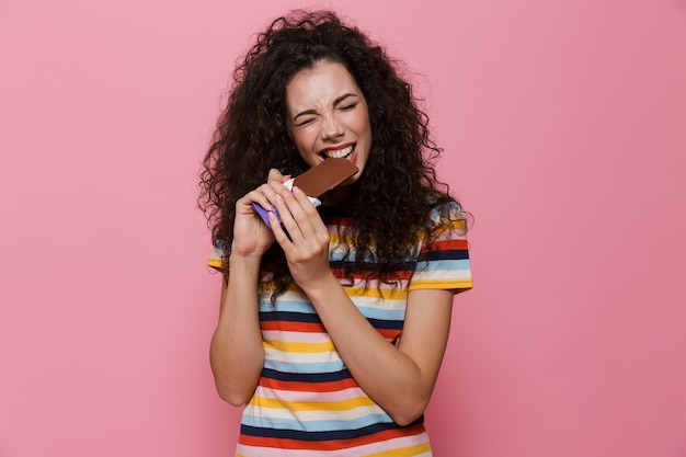 brunette woman 20s with curly hair eating chocolate bar isolated on pink