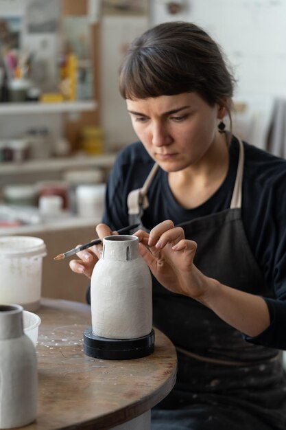 Photo brunette woman in black apron enjoys painting white handmade pottery sitting at table in workshop