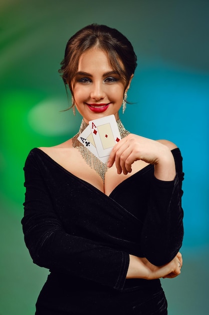 Brunette woman in black dress necklace and earrings smiling showing two playing cards posing on colo...