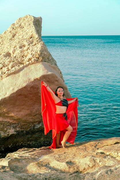 A brunette woman in a costume for oriental dances is dancing on a rock against the background of the sea with a scarf.