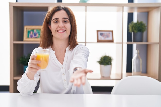 Brunette woman drinking glass of orange juice smiling cheerful offering palm hand giving assistance and acceptance