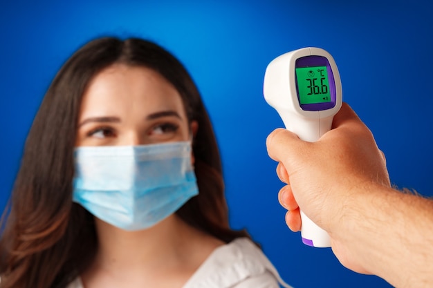 Brunette woman getting temperature screening with infrared thermometer against blue background