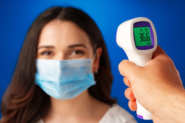 Brunette woman getting temperature screening with infrared thermometer against blue background