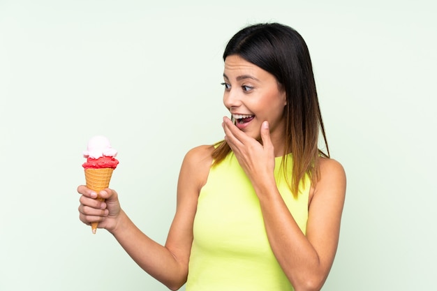 Brunette woman holding a cornet ice cream over green wall with surprise and shocked facial expression