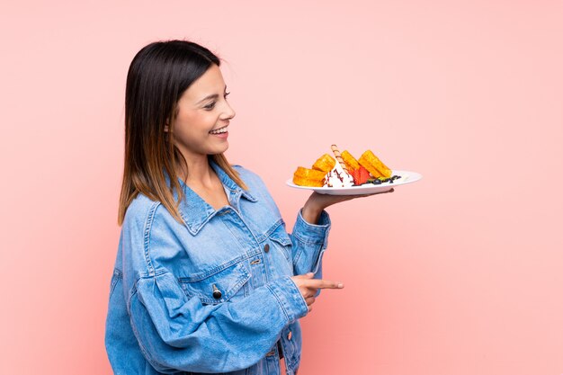 Brunette woman holding waffles over isolated pink wall pointing to the side to present a product