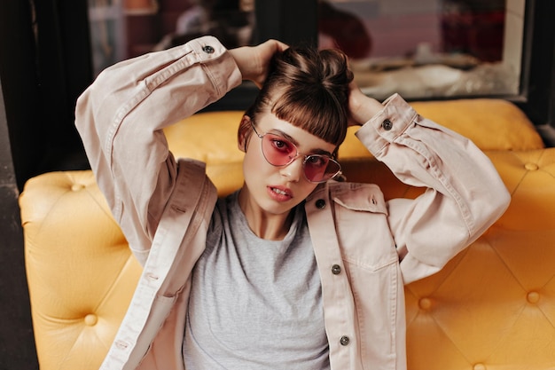 Фото Брюнетка в розовых очках сидит на желтом диване в кафе модная девушка с короткими волосами в легкой куртке позирует внутри