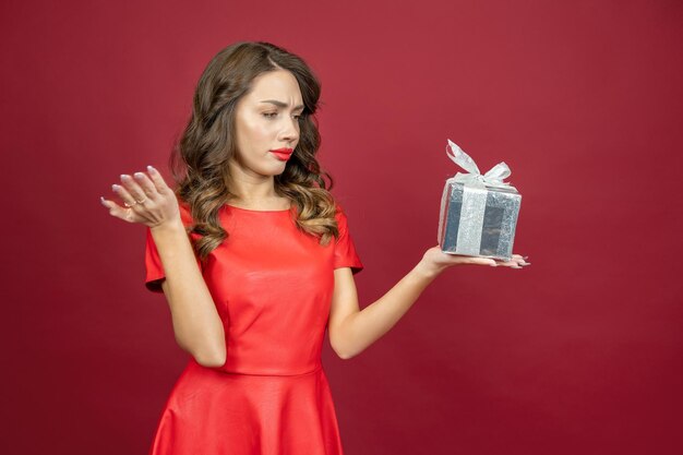 Brunette woman is disappointed with the gift in red dress on a red background