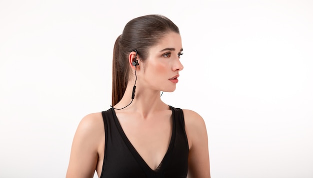 brunette woman in jogging black top listening to music on earphones posing on grey