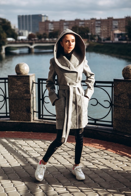 Brunette woman in a long gray coat walks along the embankment of the river