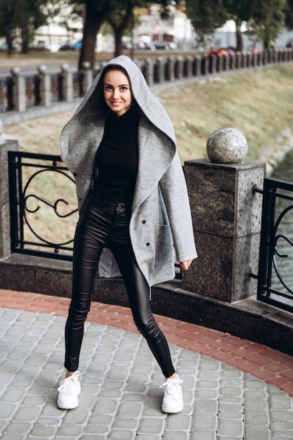 Brunette woman in a long gray coat with hoodie walks along the embankment of the river