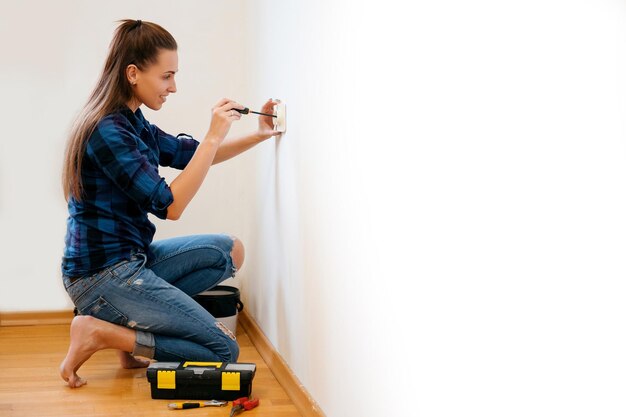 Brunette woman in plaid shirt repairs an electric socket with a\
screwdriver