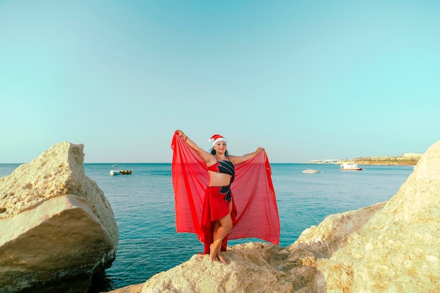 A brunette woman in a red oriental dance costume and a Santa hat is dancing on the background of the sea.