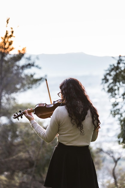 Brunette woman seen from back playing violin outside in the mountain. Vertical