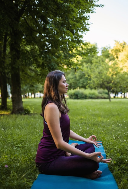 Una donna bruna si siede lateralmente e medita su una stuoia di yoga in natura