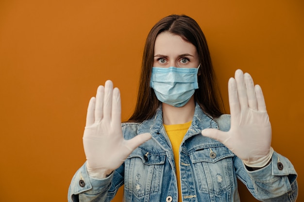 Brunette woman in sterile face mask gloves, pulls hands towards camera in stop gesture, shows limit, wears denim jacket, isolated over on brown wall. Quarantine pandemic coronavirus virus concept