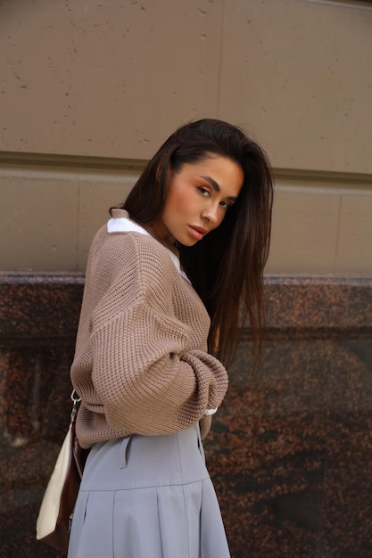 A brunette woman on the street in a brown sweater and a blue skirt next to a beige building everyday lifestyle casual