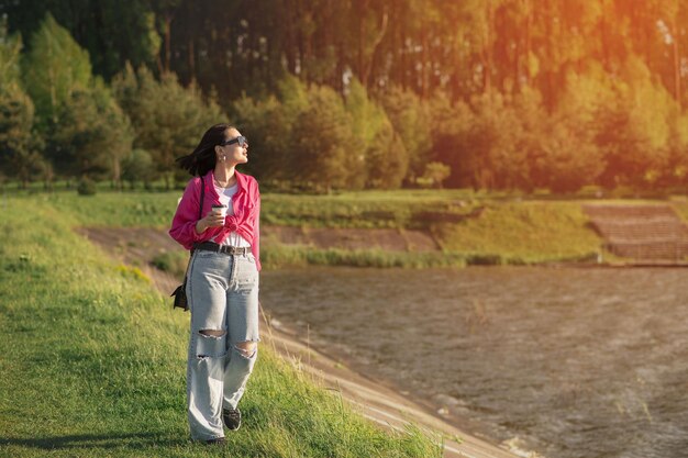 Brunette woman in sunglasses dressed in casual with cup of coffee walking around the lake shore in sunny day