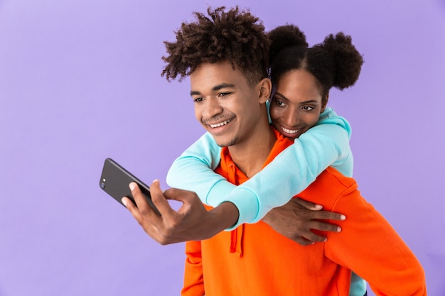 brunette woman taking selfie while hugging handsome man from back, isolated over violet wall