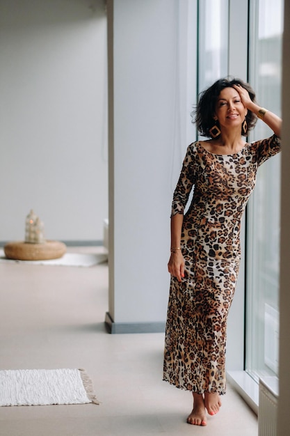 A brunette woman in a tiger dress stands near a large window.