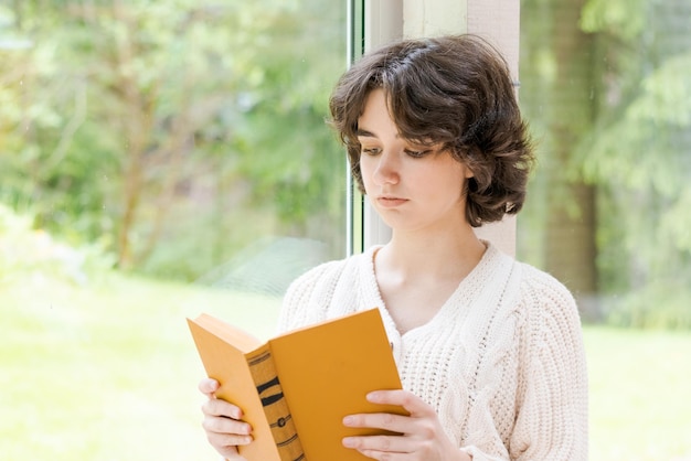 Brunette woman in warm sweater reading book sitting on room on the veranda