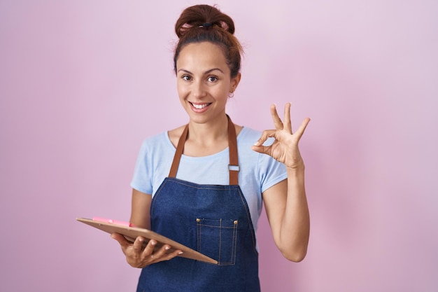 Brunette woman wearing professional waitress apron holding clipboard doing ok sign with fingers smiling friendly gesturing excellent symbol