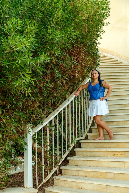 A brunette woman in a white skirt stands on a spiral staircase entwined with greenery.