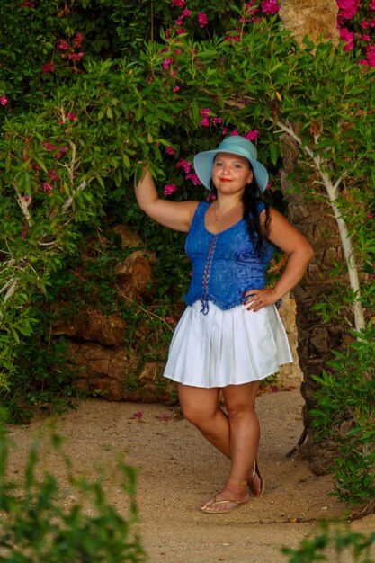 A brunette woman in a white skirt and a sun hat stands by the blooming green plants