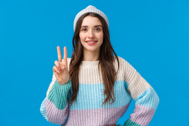 brunette woman in winter hat and sweater showing peace sign and smiling