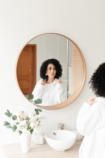 Brunette woman with African curls in a white sweater looks in the mirror