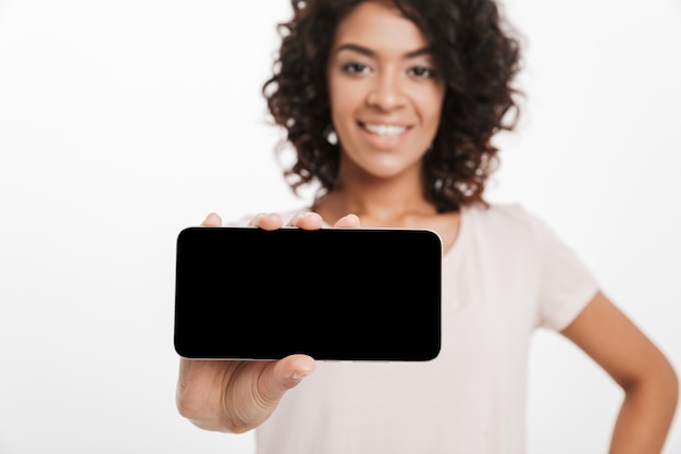 Foto donna castana con i capelli afro che fanno lo schermo commerciale e di dimostrazione del copyspace dello smartphone, isolato sopra la parete bianca