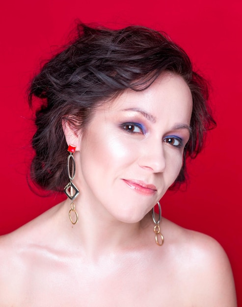 A brunette woman with beautiful makeup and bare shoulders poses in a studio on a red background in long earrings The girl looks at the camera and smiles