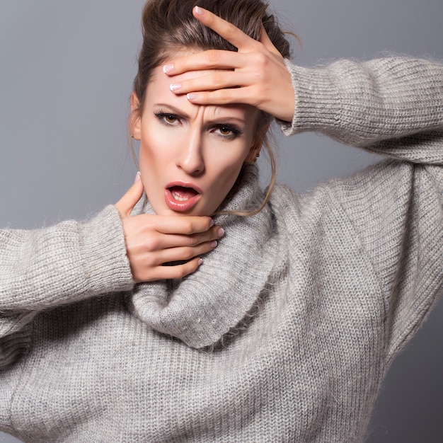 Brunette woman with bun hairstyle and neutral make up posing in grey sweater on a grey background