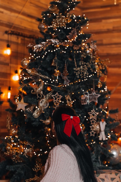 Photo a brunette woman with a red bow in her hair sitting near a christmas tree with golden decorations