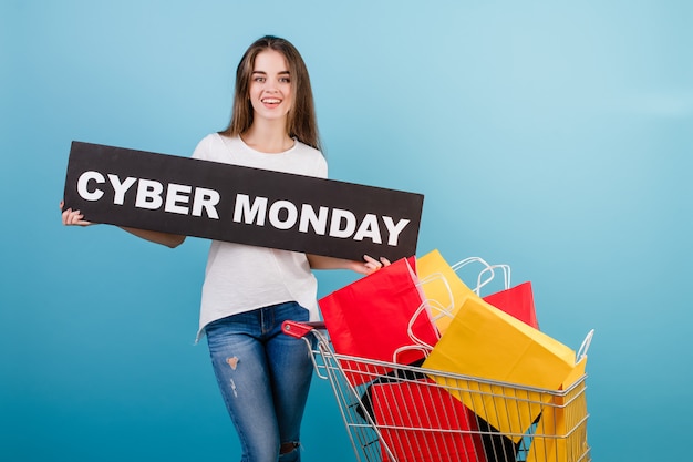 Brunette woman with shopping cart full of colorful red and yellow paper bags and cyber monday sign isolated over blue